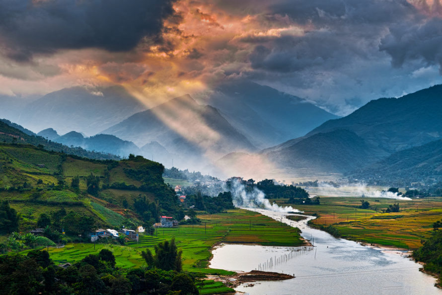 A showcase nature image featuring a deep mountain valley with a river on the foreground and a beautiful light rays penetrating the clouds and reaching the valley on the background.